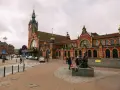 Dworzec w Gdańsku z charakterystyczną wieżą zegarową i ceglaną fasadą. Przed budynkiem rzeźba dzieci i ruch uliczny.