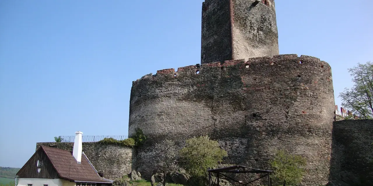 Zamek Bolków, potężna kamienna twierdza z wieżą, góruje nad zielonym wzgórzem. Obok stoi mały, drewniany domek.