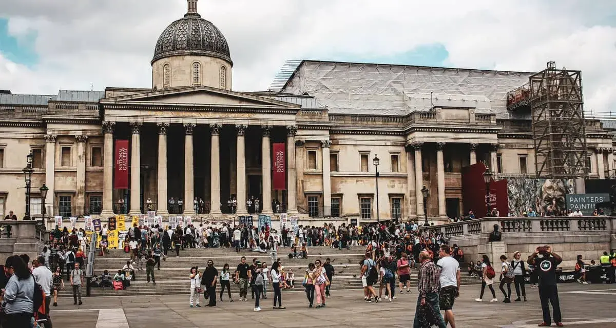 Tłum ludzi przed National Gallery w Londynie. Monumentalna fasada z kolumnami i kopułą, czerwone banery zapraszają do zwiedzania.