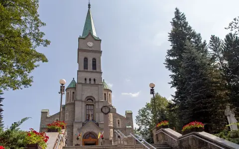 Słynny kościół w Zakopanem, z kamienną fasadą i zieloną wieżą, otoczony drzewami i ozdobiony czerwonymi kwiatami.