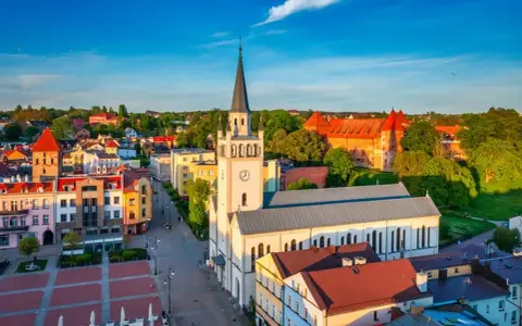 Słoneczny rynek w Bytowie z kościołem i zamkiem w tle. Kolorowe kamienice tworzą urokliwy krajobraz.