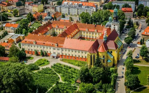 Widok z lotu ptaka na klasztor w Trzebnicy z charakterystycznymi żółtymi murami i czerwonymi dachami. Otoczony zielenią i budynkami miejskimi.