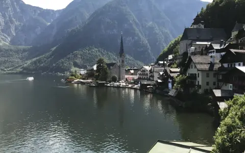 Malowniczy widok na Hallstatt, miasteczko nad jeziorem, z górami w tle. Widać kościół i domy, a na wodzie statek. To miejsce słynie z kopalni soli.