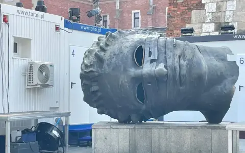 Monumentalna głowa na rynku w Krakowie, z pustymi oczodołami, stoi obok białych kontenerów.
