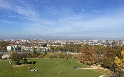 Jesienny widok na Kraków z Kopca Kraka. Ludzie odpoczywają na trawie, podziwiając panoramę miasta i kolorowe drzewa.