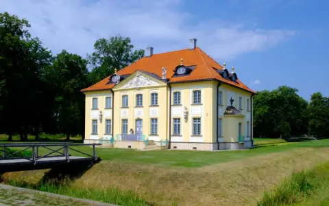 Pałac w Choroszczy, żółty, z pomarańczowym dachem, otoczony zielenią. Drewniany mostek prowadzi do posiadłości.