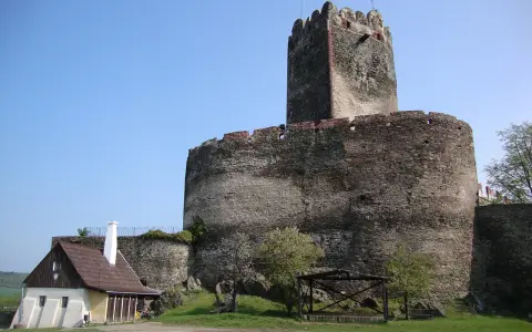 Zamek Bolków, potężna kamienna twierdza z wieżą, góruje nad zielonym wzgórzem. Obok stoi mały, drewniany domek.