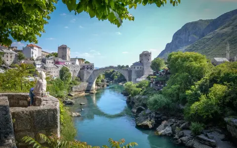 Widok na Mostar w Bośni z kamiennym mostem Stary Most nad turkusową rzeką Neretwą. W tle kamienne budynki i góry.