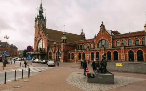 Dworzec w Gdańsku z charakterystyczną wieżą zegarową i ceglaną fasadą. Przed budynkiem rzeźba dzieci i ruch uliczny.