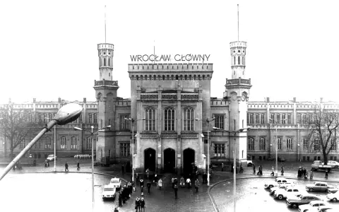 Wrocław Główny, imponujący dworzec z wieżami i zegarem, otoczony samochodami i ludźmi.