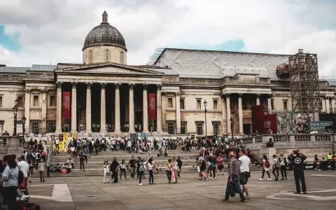 Tłum ludzi przed National Gallery w Londynie. Monumentalna fasada z kolumnami i kopułą, czerwone banery zapraszają do zwiedzania.