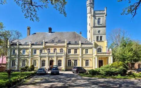Żółty pałac w Rybokartach z wieżą, otoczony zielenią.