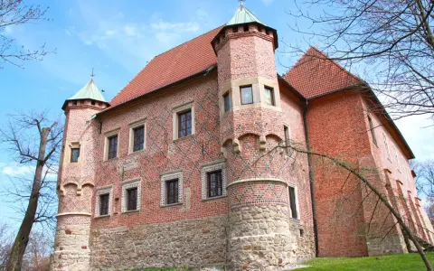 Zamek w Dębnie, ceglana twierdza z kamienną podstawą i zielonymi wieżyczkami, otoczony drzewami pod błękitnym niebem.