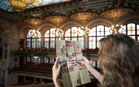 Kobieta studiuje broszurę z widokiem na wspaniałe wnętrze Pałacu Muzyki Katalońskiej, z witrażowym sufitem i rzędami siedzeń.