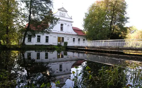 Biały kościół na wodzie w Zwierzyńcu, z odbiciem w tafli jeziora i mostkiem.