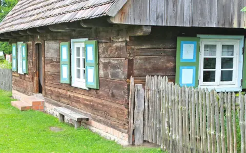 Drewniana chata z zielono-niebieskimi okiennicami, ławką i płotem. Skansen w Dziekanowicach zaprasza do podróży w czasie.