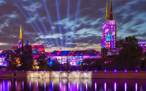 Oświetlony nocą Ostrów Tumski we Wrocławiu, z kolorowymi projekcjami na budynkach i niebieskimi snopami światła na niebie.