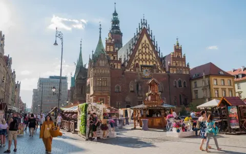 Tłum ludzi spaceruje po starym rynku we Wrocławiu, podziwiając piękną architekturę i letnie stoiska.
