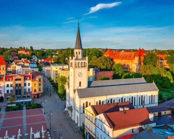 Słoneczny rynek w Bytowie z kościołem i zamkiem w tle. Kolorowe kamienice tworzą urokliwy krajobraz.
