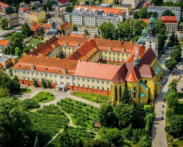 Widok z lotu ptaka na klasztor w Trzebnicy z charakterystycznymi żółtymi murami i czerwonymi dachami. Otoczony zielenią i budynkami miejskimi.