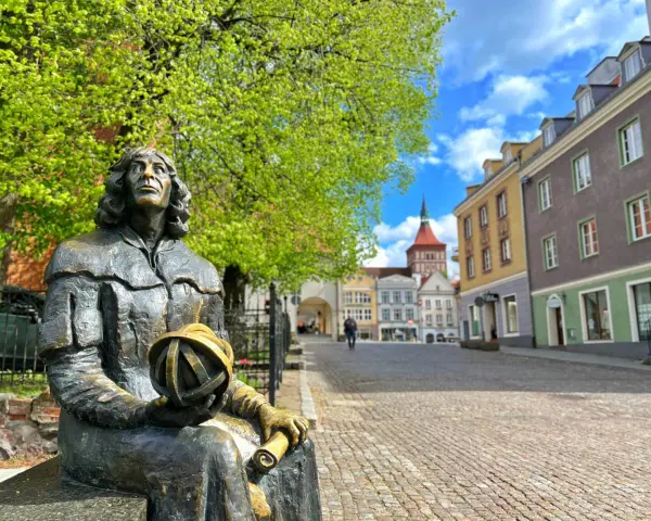 Pomnik Kopernika na olsztyn rynek, z kolorowymi kamienicami w tle i zielonymi drzewami.