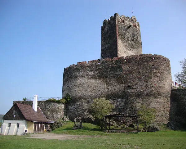 Zamek Bolków, potężna kamienna twierdza z wieżą, góruje nad zielonym wzgórzem. Obok stoi mały, drewniany domek.