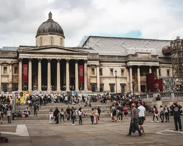 Tłum ludzi przed National Gallery w Londynie. Monumentalna fasada z kolumnami i kopułą, czerwone banery zapraszają do zwiedzania.