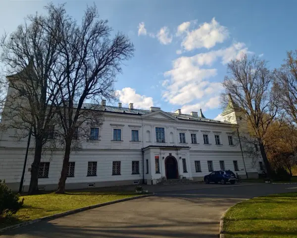 Pałac w Falentach, biała, okazała budowla z wieżyczką, otoczona drzewami o nagich gałęziach, pod błękitnym niebem z chmurami.