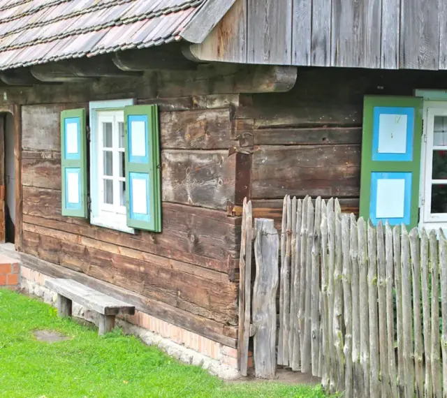 Drewniana chata z zielono-niebieskimi okiennicami, ławką i płotem. Skansen w Dziekanowicach zaprasza do podróży w czasie.