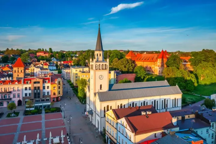 Słoneczny rynek w Bytowie z kościołem i zamkiem w tle. Kolorowe kamienice tworzą urokliwy krajobraz.