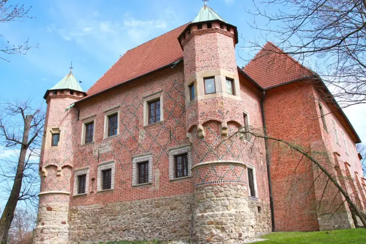 Zamek w Dębnie, ceglana twierdza z kamienną podstawą i zielonymi wieżyczkami, otoczony drzewami pod błękitnym niebem.