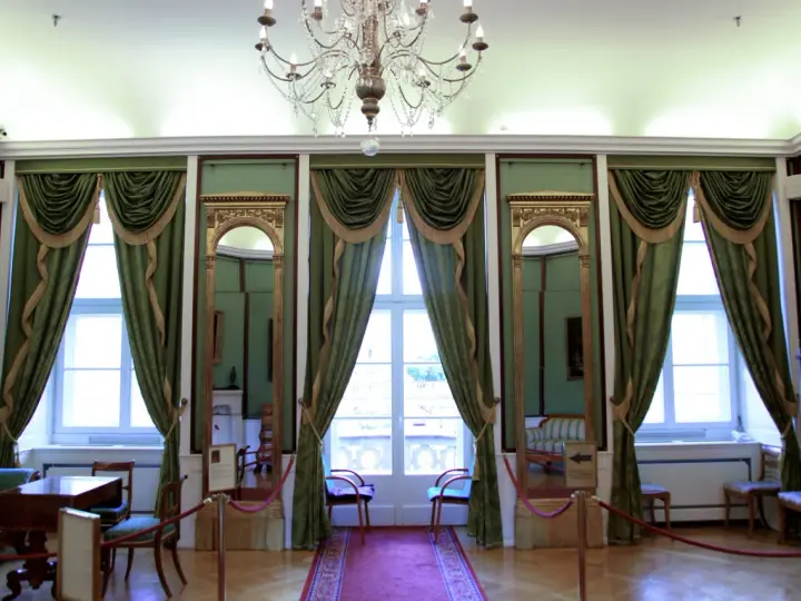 Wnętrze pałacu królewskiego we Wrocławiu. Eleganckie zielone zasłony, złote lustra i kryształowy żyrandol tworzą atmosferę dawnej świetności.