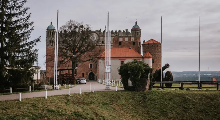 Zamek Golub-Dobrzyń zaprasza na zwiedzanie. Potężna twierdza z armatą na dziedzińcu, otoczona zielenią i drzewami.