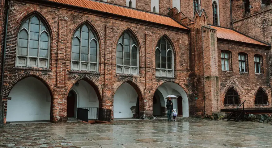 Zamek w Kwidzynie, z ceglanymi arkadami i gotyckimi oknami, skrywa tajemnice. Dwoje ludzi z parasolem spaceruje po mokrym dziedzińcu.