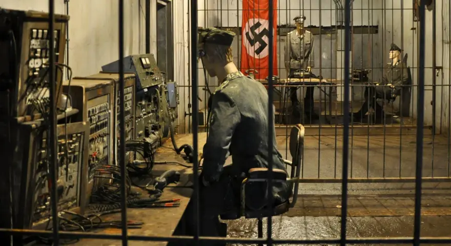 Wnętrze mamerki bunkrów z manekinami żołnierzy i niemiecką flagą. Na pierwszym planie manekin przy konsoli, w tle dwaj żołnierze.