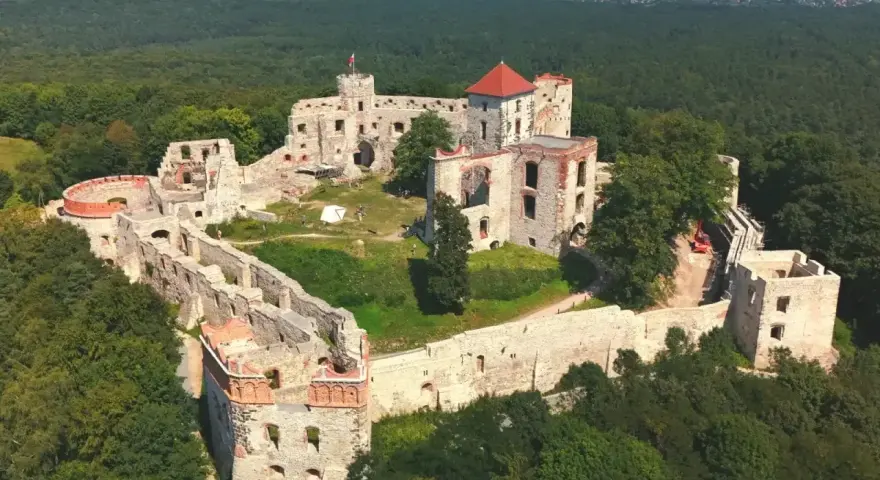 Ruiny zamku Tenczyn na wzgórzu, otoczone lasem. Widać mury obronne, wieże i pozostałości budynków.