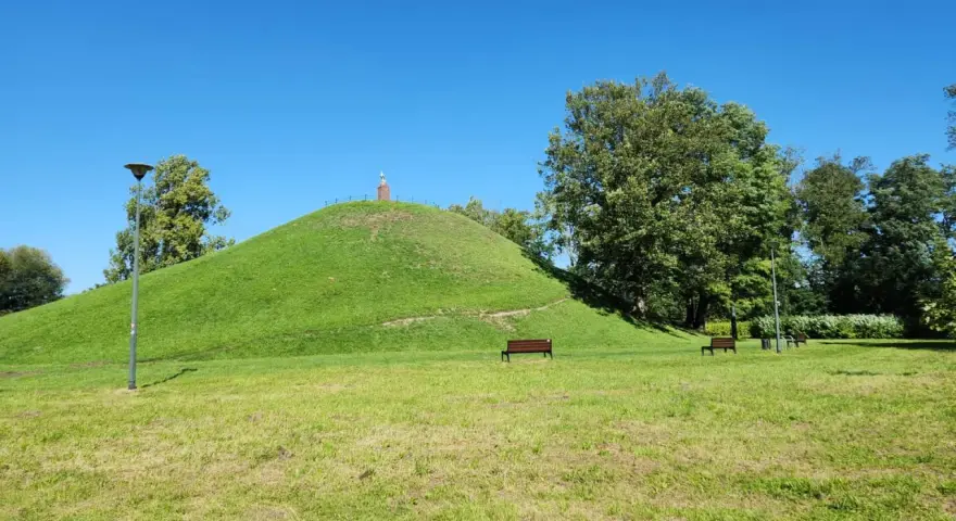 Zielony kopiec Wandy z pomnikiem na szczycie, otoczony drzewami i ławkami w parku.