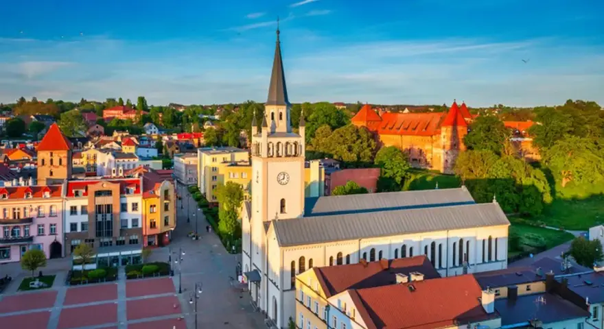 Słoneczny rynek w Bytowie z kościołem i zamkiem w tle. Kolorowe kamienice tworzą urokliwy krajobraz.