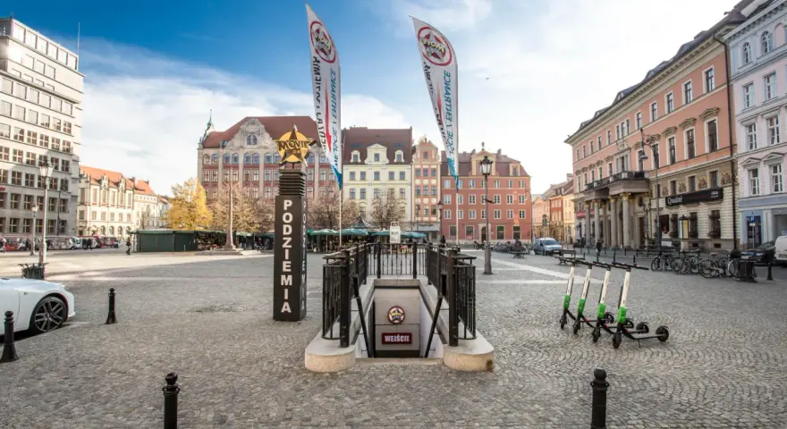 Wejście do podziemi z napisem "Movie Gate Wrocław" na placu w otoczeniu zabytkowych kamienic.
