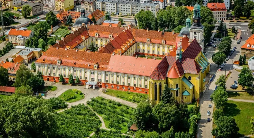 Widok z lotu ptaka na klasztor w Trzebnicy z charakterystycznymi żółtymi murami i czerwonymi dachami. Otoczony zielenią i budynkami miejskimi.