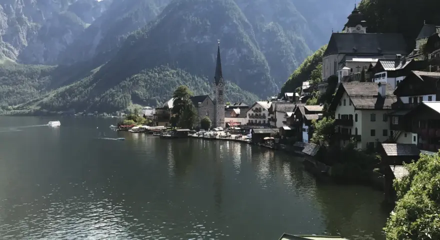 Malowniczy widok na Hallstatt, miasteczko nad jeziorem, z górami w tle. Widać kościół i domy, a na wodzie statek. To miejsce słynie z kopalni soli.