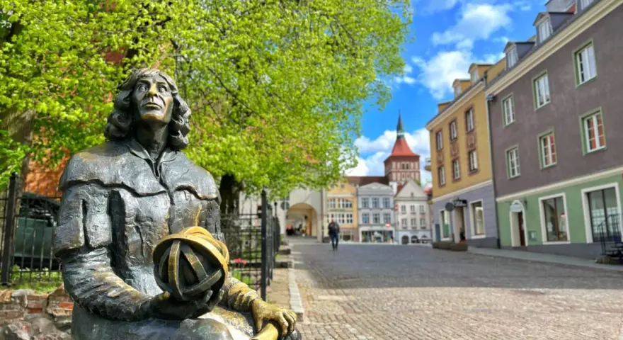 Pomnik Kopernika na olsztyn rynek, z kolorowymi kamienicami w tle i zielonymi drzewami.