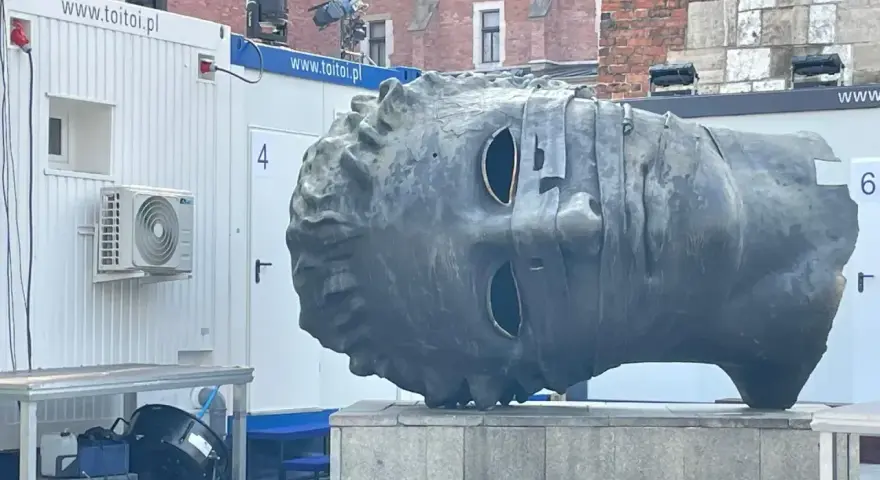 Monumentalna głowa na rynku w Krakowie, z pustymi oczodołami, stoi obok białych kontenerów.