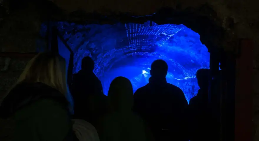 Grupa turystów wchodzi do tunelu oświetlonego niebieskim światłem. To część podziemnej trasy turystycznej w dawnej kopalni uranu Kowary.