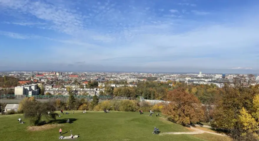 Jesienny widok na Kraków z Kopca Kraka. Ludzie odpoczywają na trawie, podziwiając panoramę miasta i kolorowe drzewa.