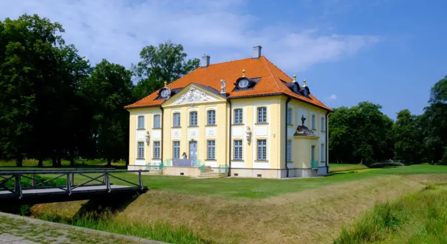 Pałac w Choroszczy, żółty, z pomarańczowym dachem, otoczony zielenią. Drewniany mostek prowadzi do posiadłości.