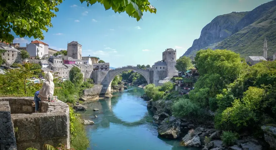 Widok na Mostar w Bośni z kamiennym mostem Stary Most nad turkusową rzeką Neretwą. W tle kamienne budynki i góry.