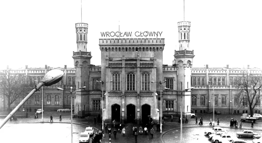 Wrocław Główny, imponujący dworzec z wieżami i zegarem, otoczony samochodami i ludźmi.