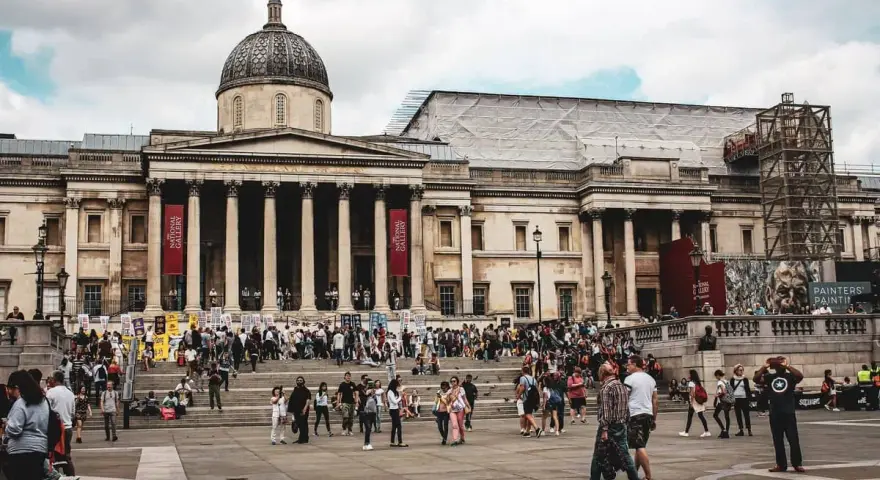 Tłum ludzi przed National Gallery w Londynie. Monumentalna fasada z kolumnami i kopułą, czerwone banery zapraszają do zwiedzania.