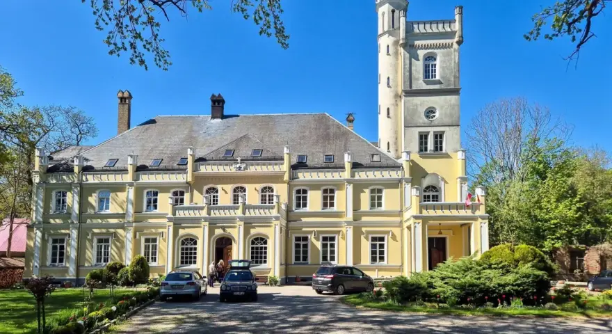 Żółty pałac w Rybokartach z wieżą, otoczony zielenią.